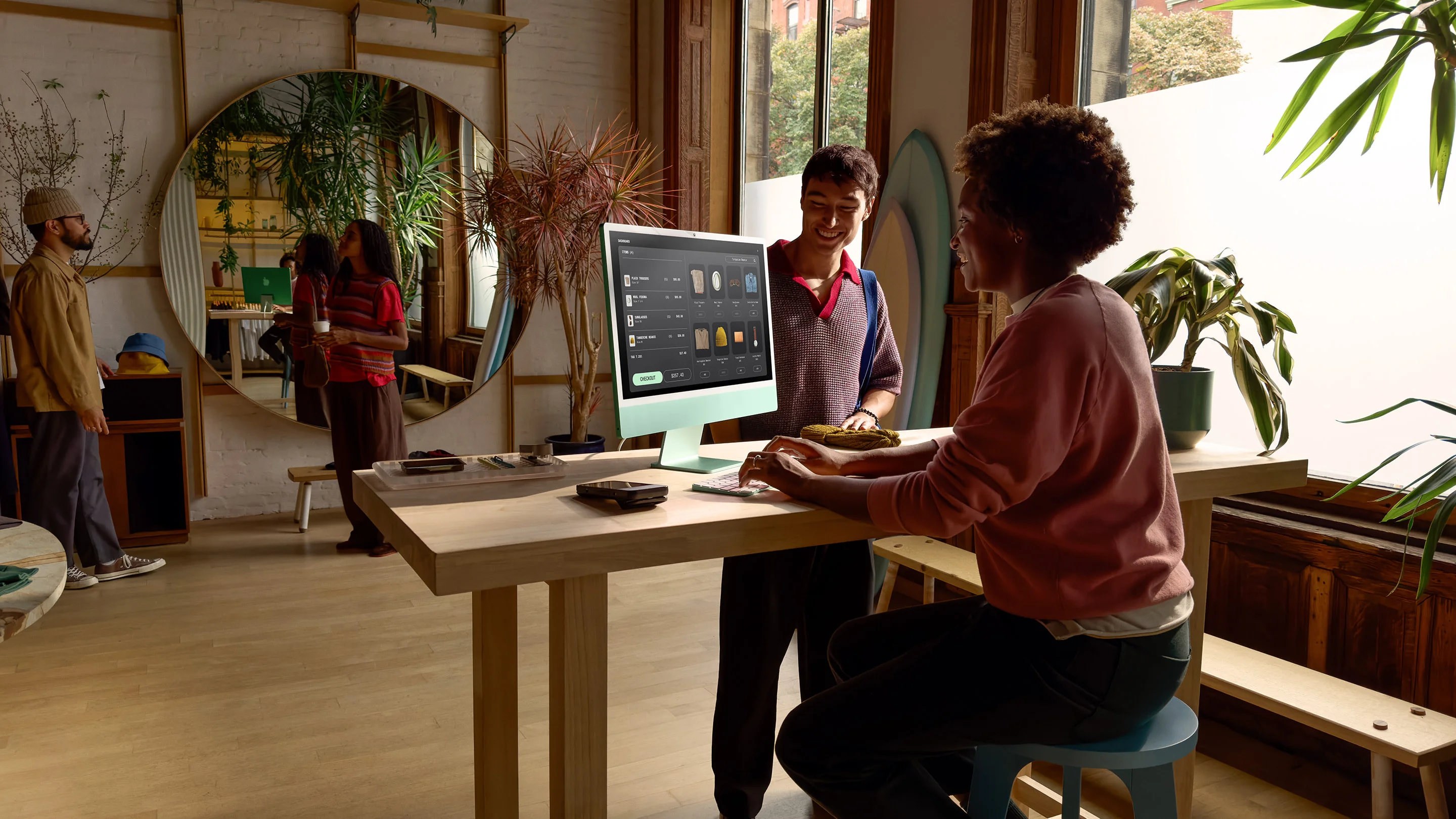 A modern store setting with two people using an Apple iMac at a high wooden table. One person is seated, interacting with the iMac, which displays a product catalogue, while the other person stands, smiling his hand on a wollen hat placed on the table. The store has large windows, potted plants, and a surfboard leaning against the wall, with additional people and decor visible in the reflection of a large circular mirror.