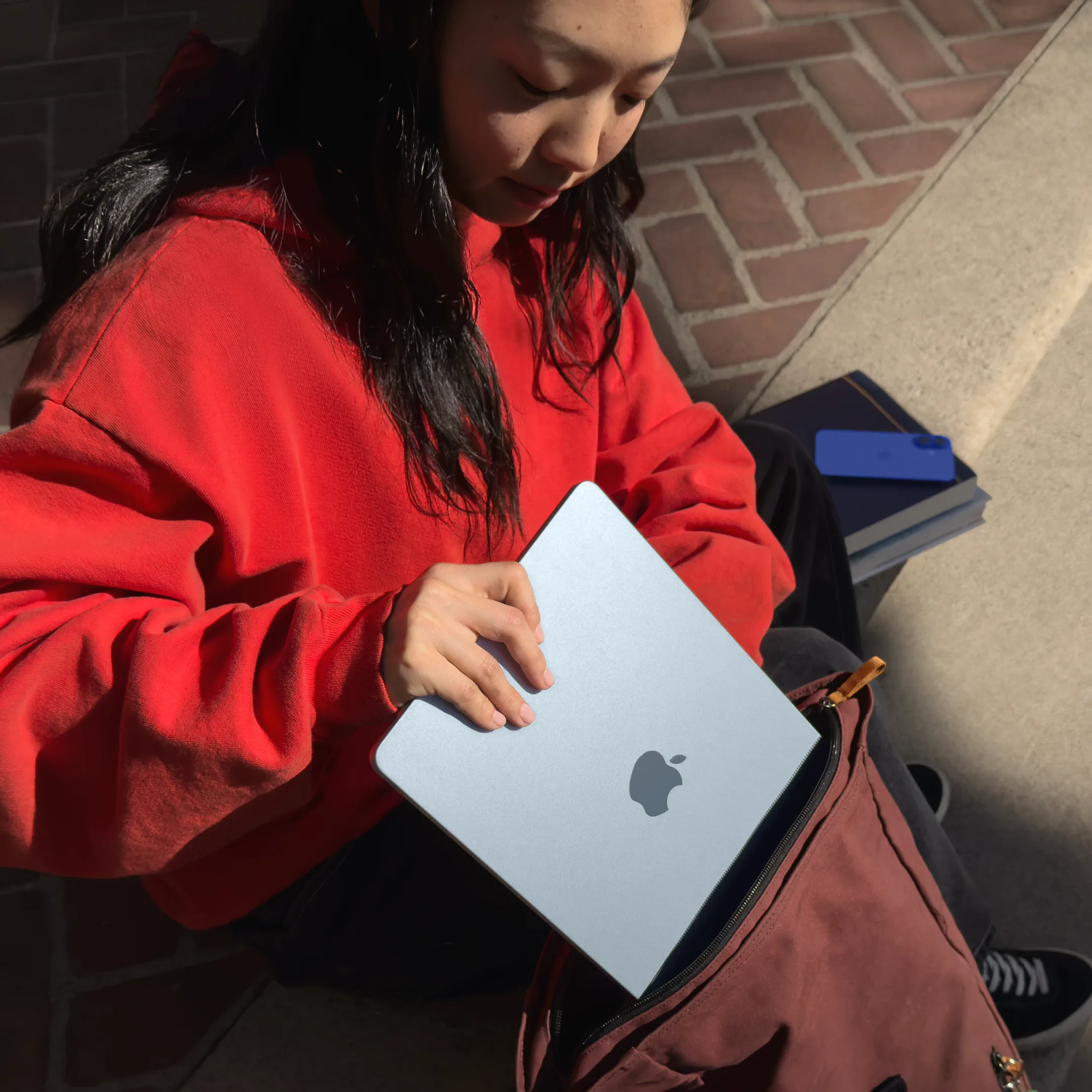 A person in a red hoodie placing a Sky Blue MacBook Air into a maroon backpack, sitting outdoors with books and a smartphone nearby.