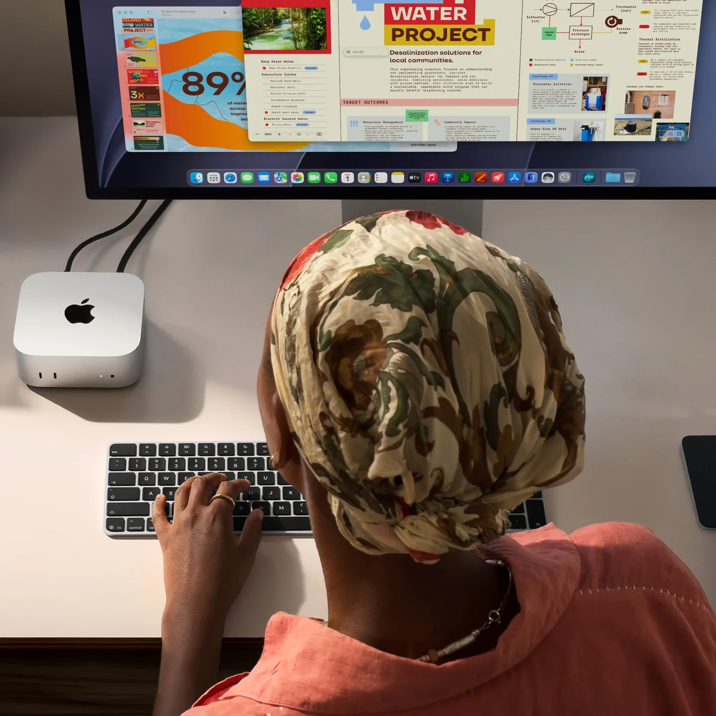 A person at a white desk working on a Mac mini connected to a Studio Display, hands on a Magic Keyboard and Magic Trackpad, with a stack of plates and a tape dispenser at the sides