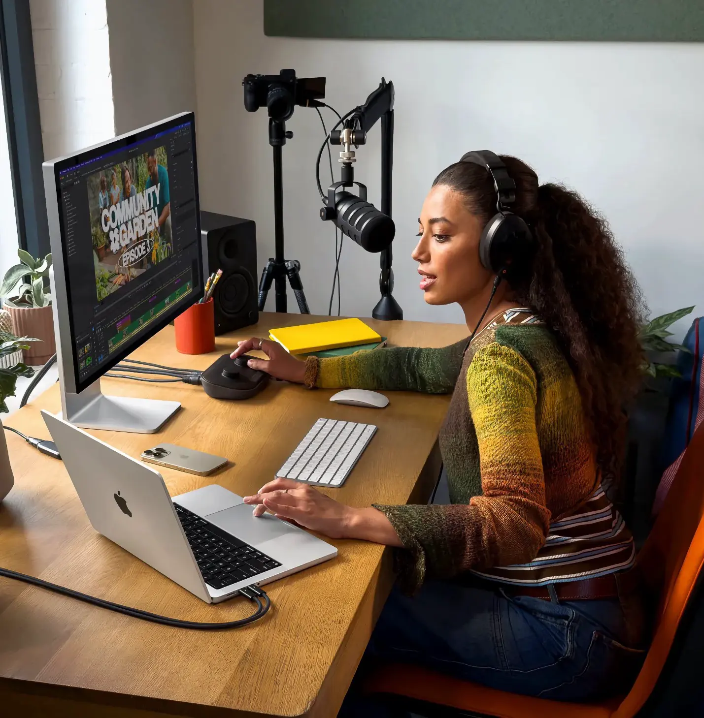 A modern store setting with two people at a high wooden table. One person is seated, working on a Mac which displays a product catalogue, while the other person stands smiling. The store has large windows, potted plants, and a surfboard leaning against the wall, with additional people and decor visible in the reflection of a large circular mirror.