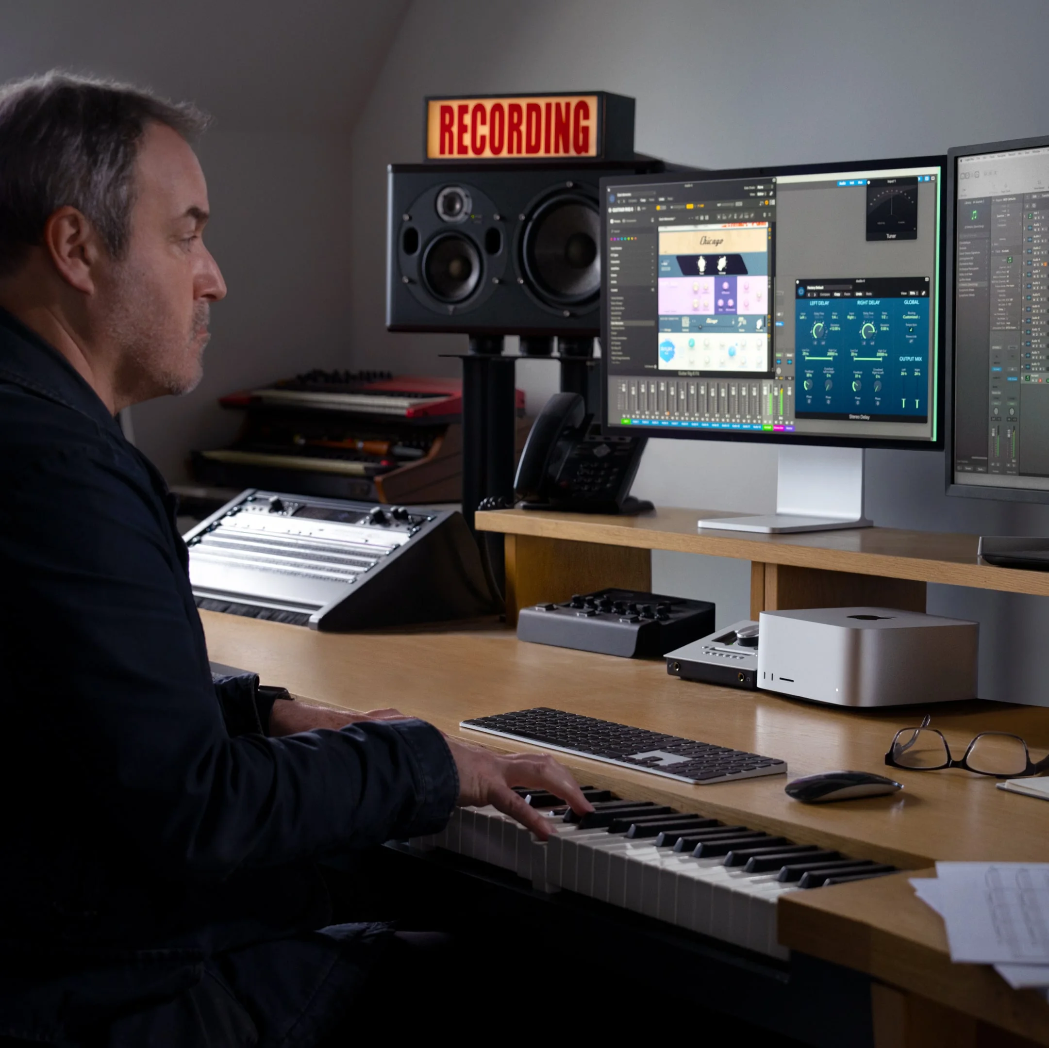 A music producer working in a modern studio, playing a MIDI keyboard and using a dual-monitor setup with music production software on a Mac Studio. The workspace includes studio monitors, a recording sign, and various music equipment.